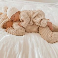 two babies are laying on a bed together, one is wearing a sweater and the other has a hat