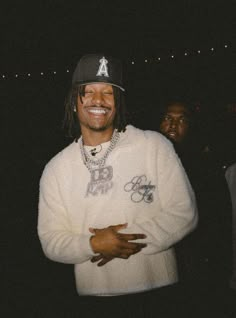 a man in a white sweatshirt and black hat standing with his arms crossed, smiling at the camera