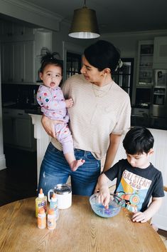 a woman holding a baby while standing next to two other children at a kitchen table