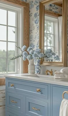 a blue and white bathroom with flowers on the counter top, gold framed mirror above it