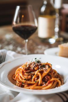 a white plate topped with pasta and sauce next to a glass of wine on a table