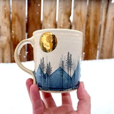 a hand holding a coffee cup with trees on it in front of a wooden fence