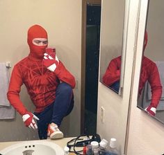 a man in a red spider suit is sitting on a bathroom counter looking at his cell phone