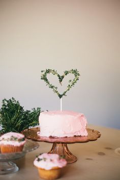 a pink cake sitting on top of a wooden table next to cupcakes and greenery