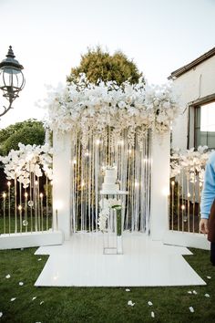 a white wedding ceremony setup with flowers and candles on the grass in front of a building