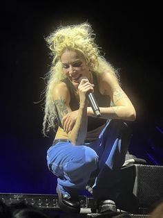 a woman sitting on top of a stage with a microphone in her hand and wearing blue pants