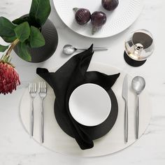 a place setting with black and white plates, silverware and flowers on a marble table