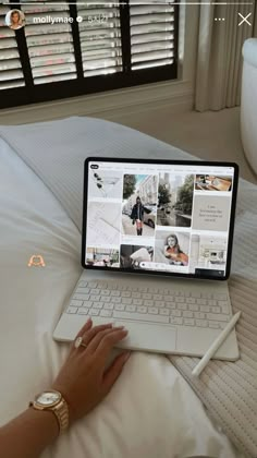 a person using a laptop computer on a bed with white sheets and windows in the background