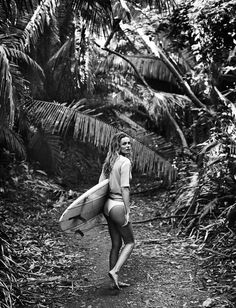 black and white photograph of a woman carrying a surfboard on a path surrounded by palm trees