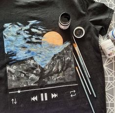 a t - shirt with an image of mountains and clouds painted on it next to paintbrushes