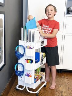 a young boy standing next to a white cart filled with toys and supplies in a kitchen