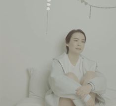 a woman sitting on top of a bed next to a white wall with lights hanging from the ceiling