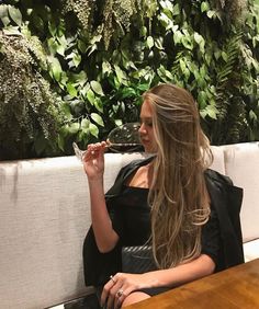 a woman sitting at a table with a glass of wine in her hand and greenery on the wall behind her