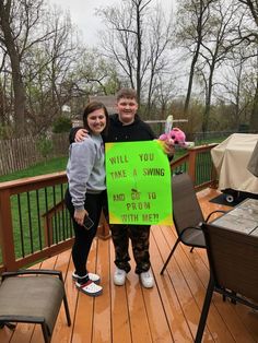 a man and woman standing on a deck holding up a sign that says, will you take a swing and go to prom with me?