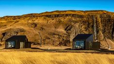 three small black cabins sitting on top of a dry grass covered field next to a mountain