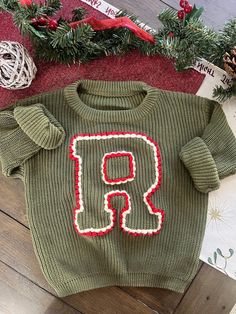 a green sweater with the letter r on it sitting next to christmas decorations and yarn