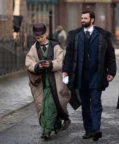 two men walking down the street one is looking at his cell phone and the other is wearing a coat