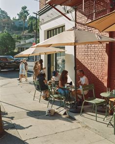 people are sitting at tables outside on the sidewalk with an umbrella over them and a dog laying under it