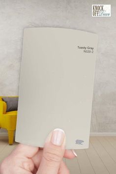 a hand holding up a gray paint swatch in front of a yellow chair and wall