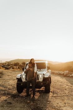 a man and woman standing next to a white truck in the middle of a dirt road