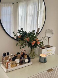 a white dresser topped with lots of bottles and flowers in front of a round mirror