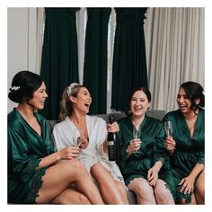 four women in green robes sitting on a couch drinking wine and laughing at each other