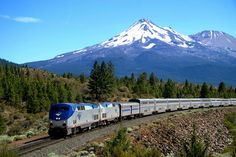 a train traveling past a snow covered mountain on the tracks in front of trees and bushes