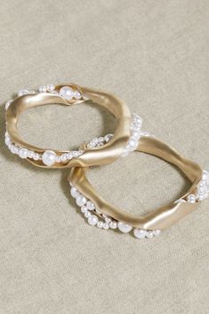 two gold rings with pearls on them sitting on top of a beige cloth covered surface