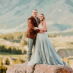 a bride and groom standing on top of a rock in front of the mountains with their arms around each other