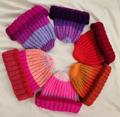 six knitted hats arranged in a circle on top of a white sheet with the colors of pink, purple, orange and red