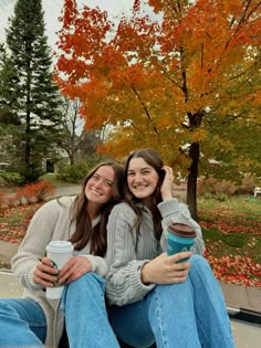 two young women sitting on the ground with coffee cups in their hands and smiling at the camera