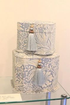 three tiered blue and white cake with tassels on glass shelf in front of wall