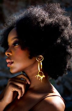 a woman with an afro is wearing gold earrings and a brown dress, looking off to the side