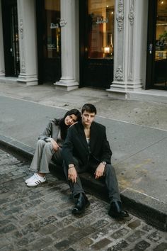 a man and woman sitting on the ground next to each other in front of a building