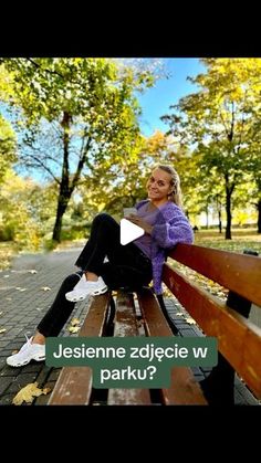 a woman sitting on top of a wooden bench in a park next to trees and leaves