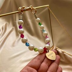 a hand holding a bracelet with pearls and charms on it's end, in front of a gold box