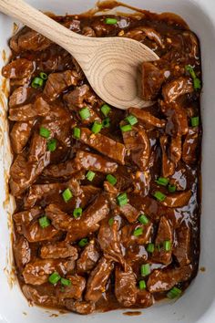 a wooden spoon in a white casserole dish filled with beef and sauce, garnished with green onions