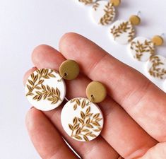 two white and gold earrings with leaves on them are being held by someone's hand