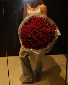 a woman standing in front of a mirror holding a bouquet of red roses and looking at her cell phone