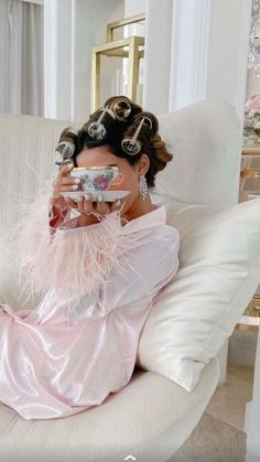 a woman sitting on top of a white couch holding a cup in her hand and wearing a pink dress