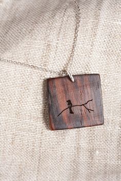 a bird is sitting on a tree branch in a wooden square pendant that hangs from a silver chain