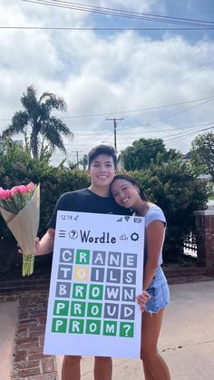 two people standing next to each other holding flowers and a sign with words on it