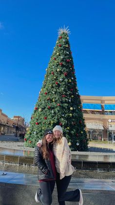 two women pose in front of a christmas tree at the center of an outdoor plaza