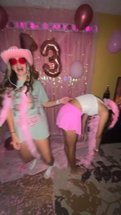 two women in pink and white outfits posing for the camera with balloons on the wall behind them