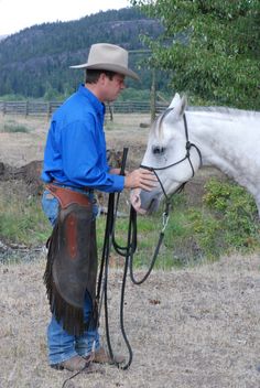 a man in blue shirt and cowboy hat standing next to white horse with bridle