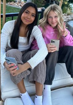 two young women sitting on a white couch holding cell phones and looking at the camera