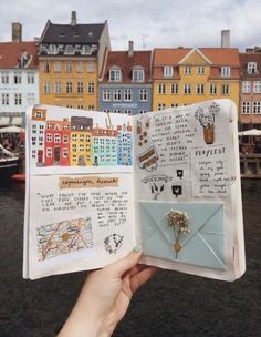 someone is holding up an open book in front of some buildings and boats on the water