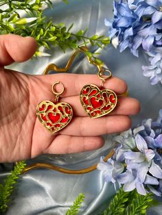 a person holding two heart shaped earrings in their hand next to blue flowers and greenery