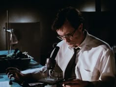 a man wearing glasses and a tie is looking at something on a table with a microscope in front of him