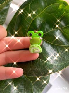 a hand holding a green frog brooch on top of a leafy plant in front of a window